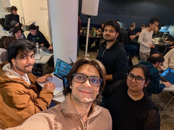 A group of people are gathered in an indoor space, with several individuals working on laptops displaying code while others pose for a selfie.