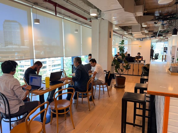 A group of people are working on laptops in a bright, modern co-working space with large windows and an industrial-style ceiling.