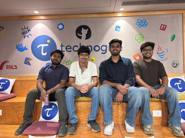 Four young men are sitting on a wooden tiered bench in front of a wall decorated with various technology logos and the company name 'technogise'.