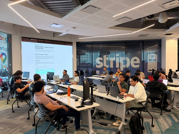 A group of people are gathered in a modern coworking space, working on laptops during what appears to be a hackathon event.