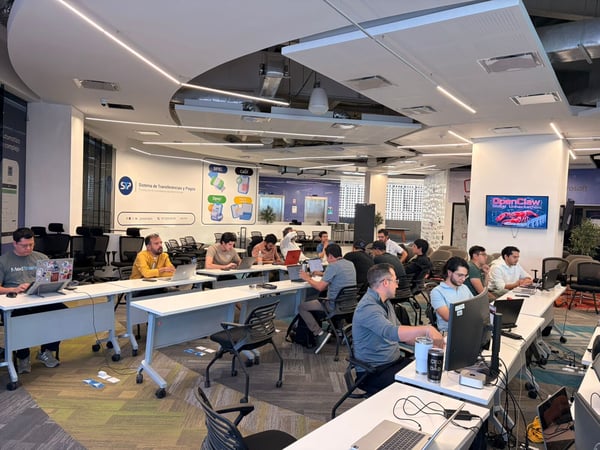 A group of men are working on laptops at long white tables in a modern, open-plan office space during a hackathon event.