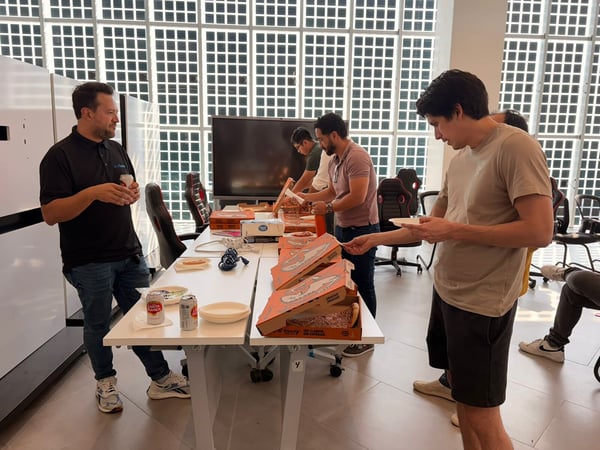 A group of men are gathered around a table in an office setting, sharing several boxes of Little Caesars pizza and drinks.