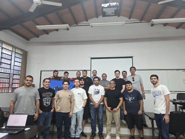 A group of eighteen men are posing for a group photograph in a classroom or workshop setting, standing in front of a whiteboard and projection screen.