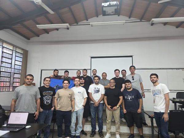 A group of about 18 men are posing for a group photo in a classroom or workshop setting, standing in front of a white screen and a whiteboard.