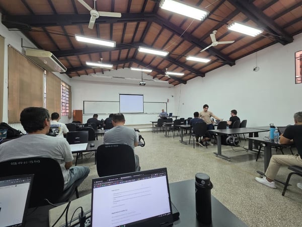 A wide-angle shot of a classroom or workshop where several people are seated at desks with open laptops, facing a presenter at the front of the room.