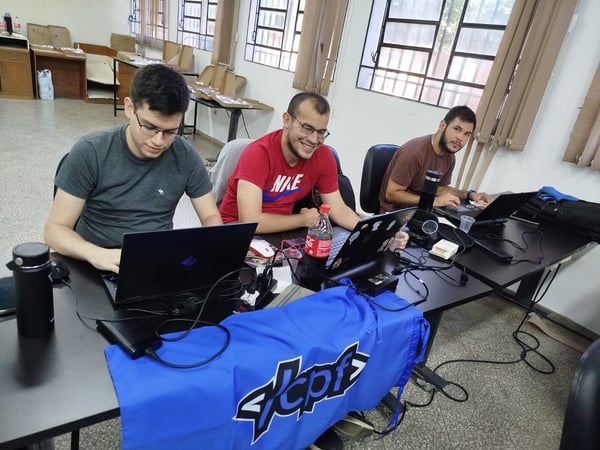 Three young men are sitting at a long table in a classroom or lab, focused on their laptops during what appears to be a coding competition or hackathon.