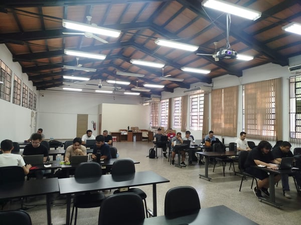 A group of people are seated at tables in a large room with a high wooden ceiling, all focused on their laptops in what appears to be a study session or workshop.