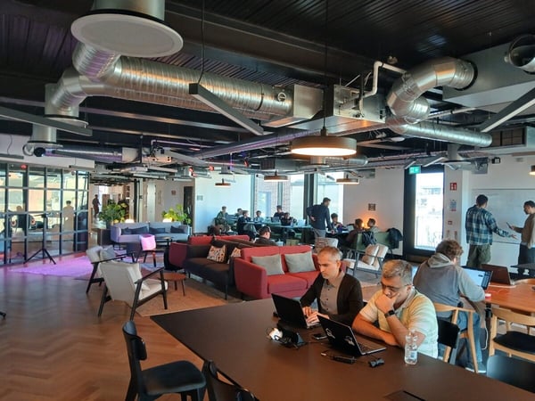 A wide shot of a modern, industrial-style co-working space with several people working on laptops at tables and in lounge areas.