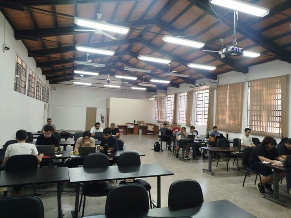 A wide-angle photograph of a large, indoor room with a high wooden truss ceiling where several people are seated at tables, most of them working on laptops.