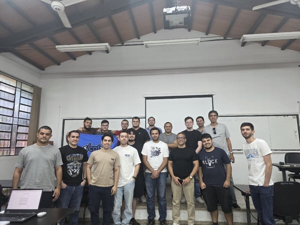 A group of eighteen men posing for a group photograph in a classroom or lecture hall, standing in front of a white projector screen and a whiteboard.