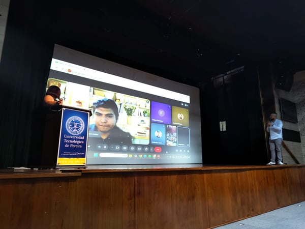 A photograph of a presentation in an auditorium, featuring a large screen displaying a Google Meet video call and a podium with the logo of the Universidad Tecnológica de Pereira.