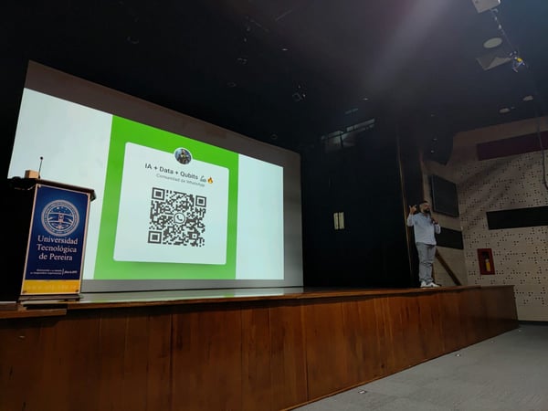 A man is giving a presentation on a stage in an auditorium, with a large screen behind him displaying a QR code for a WhatsApp community.