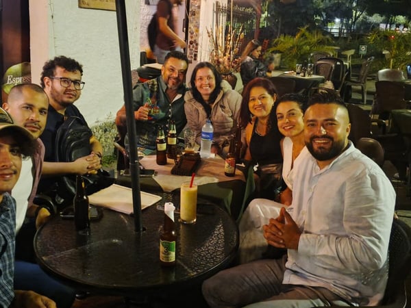 A group of eight people are smiling and posing for a photograph while sitting at outdoor tables at a restaurant or cafe during the night.