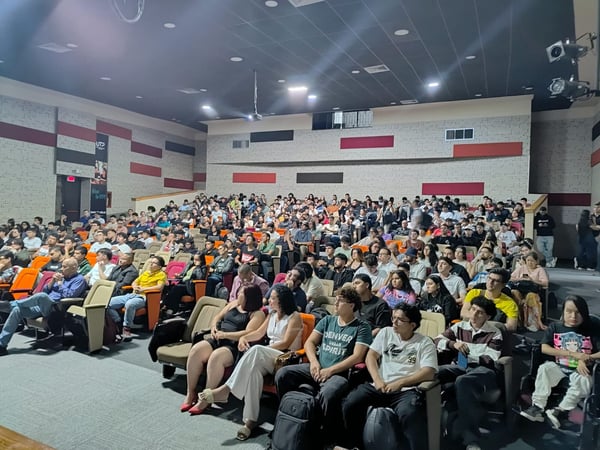 A wide-angle photograph showing a large audience of mostly young adults seated in a modern auditorium with tiered seating and acoustic wall panels.