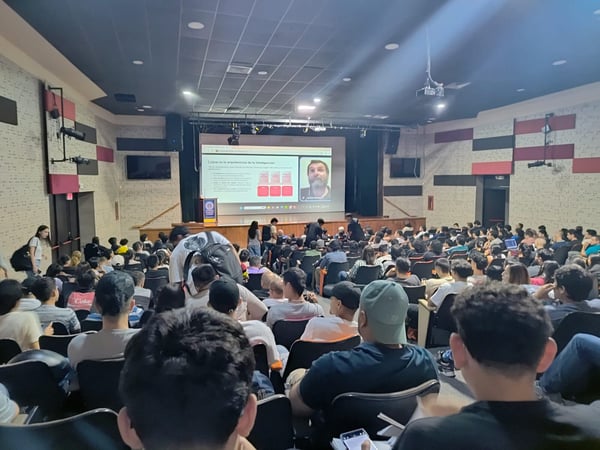 A wide-angle photograph of a large audience seated in a lecture hall, watching a presentation on a big screen that features a speaker and Spanish text.