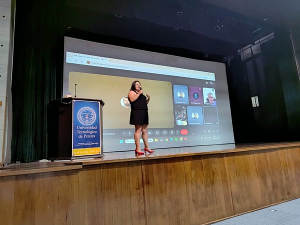 A woman stands on a wooden stage giving a presentation in front of a large screen displaying a Google Meet video call. To her left is a podium featuring the logo and name of the Universidad Tecnológica de Pereira.