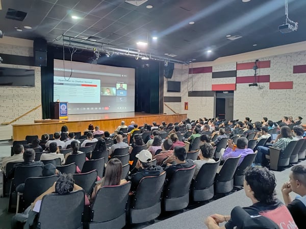 An indoor photograph of an audience seated in an auditorium, watching a presentation on a large screen about OpenAI and ChatGPT.