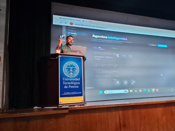 A man is giving a presentation on a stage, standing behind a podium with the Universidad Tecnológica de Pereira logo, while a large screen behind him displays a Google Meet session titled 'Agentes Inteligentes'.