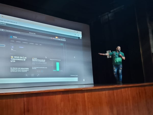 A man stands on a stage giving a presentation, gesturing toward a large projection screen displaying a dashboard while a podium with a university logo stands to the left.