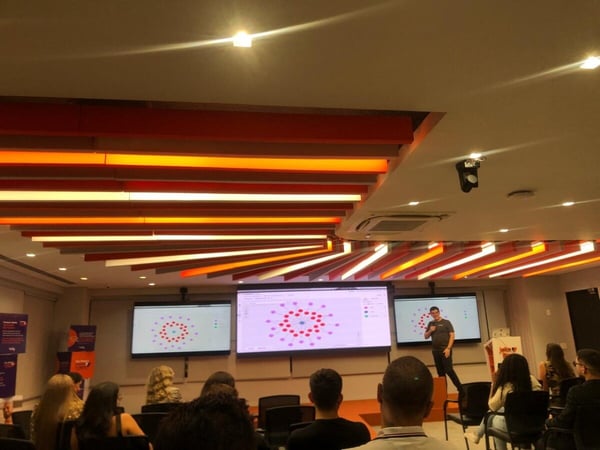 A man is delivering a presentation in a modern conference room with an audience and three large screens displaying network diagrams.
