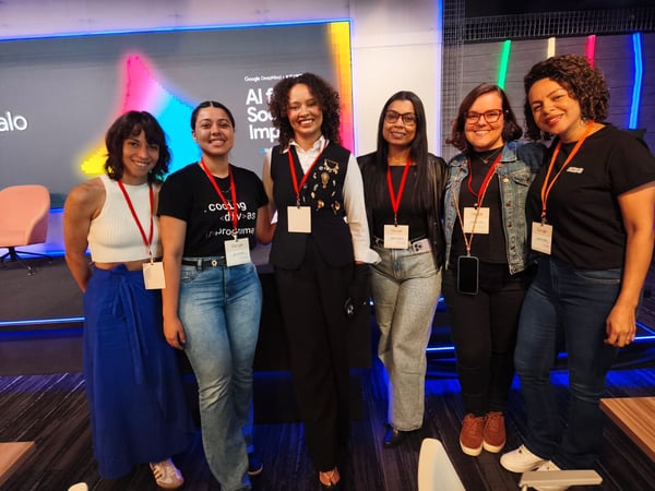 A group of six women posing for a photo at a Google DeepMind event, standing in front of a screen with the text 'AI for Social Impact'.
