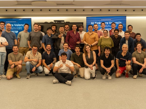 A group photo of approximately 33 people at an 'AI Tinkerers Valencia' meetup, posing in an indoor event space with digital screens and a whiteboard.