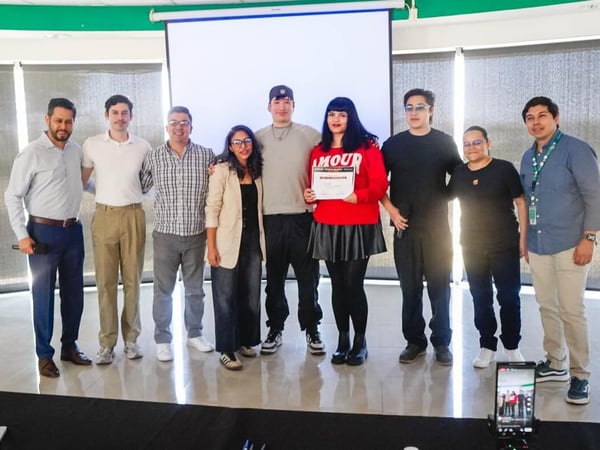 A group of nine people posing for a photograph indoors in front of a projector screen, with one woman in a red sweater holding a certificate.