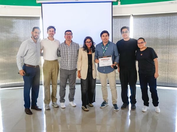 A group of seven people posing for a photo in an indoor space, with one person in the center holding a certificate in front of a projection screen.