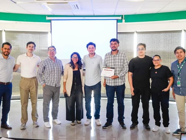 A group of nine people posing for a group photo in an indoor setting, standing in front of a large white projector screen.