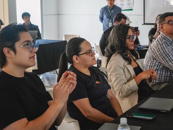 A group of people are seated at a table covered with a black cloth, attentively watching a presentation in an indoor setting.