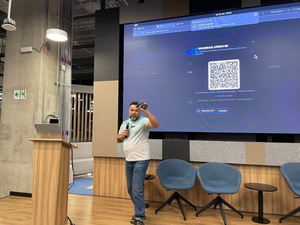 A man is giving a presentation in a modern office or event space, standing next to a podium in front of a large screen displaying a QR code.