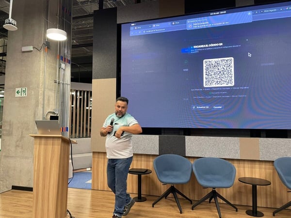 A man is giving a presentation in a modern office setting, standing next to a large screen displaying a QR code for a WhatsApp connection.