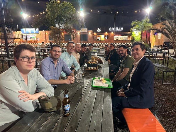 A group of eleven people are sitting around a long wooden picnic table at an outdoor food court at night, posing for a photo.