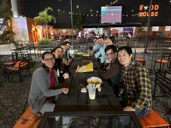 A group of friends or colleagues are gathered around a long outdoor table at night, posing for a photo at a food court area with a 'FOOD HOUSE' sign in the background.