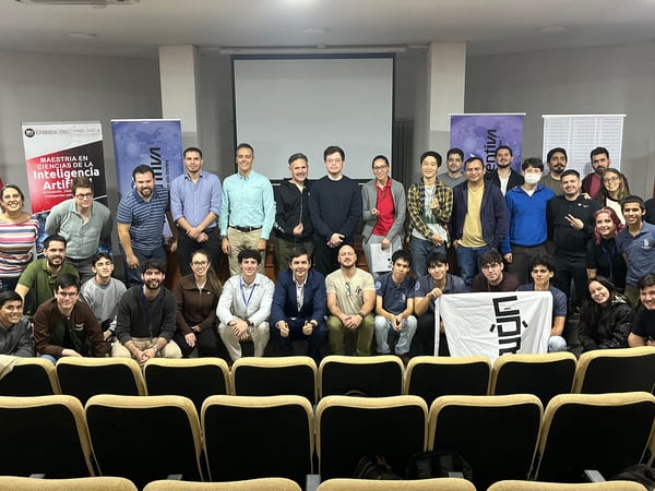 A group photo of approximately 32 people, mostly young adults, posing in an auditorium in front of a projection screen and several event banners.