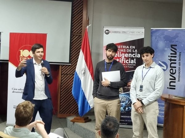 A man in a blue suit is giving a presentation at an event, standing in front of banners and a Paraguayan flag, with two other men beside him.