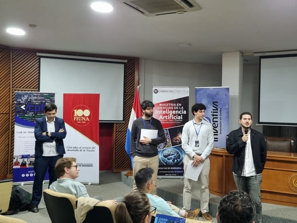 A group of men are giving a presentation in an auditorium, with one man speaking into a microphone in front of several informational banners.