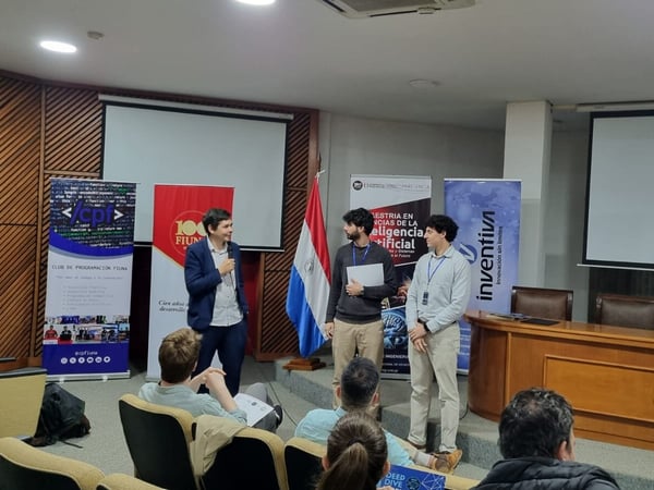 A group of men are on a stage in an auditorium giving a presentation, with banners and the flag of Paraguay behind them.