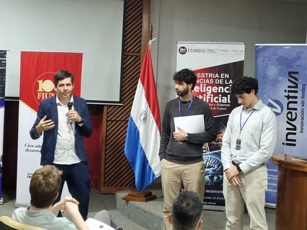A man in a blue blazer speaks into a microphone during a presentation, flanked by two other men in front of several informational banners and the Paraguayan flag.