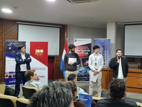 A group of men are standing at the front of a lecture hall during a presentation, with one man speaking into a microphone. Banners related to the Faculty of Engineering (FIUNA) and Artificial Intelligence are visible in the background.