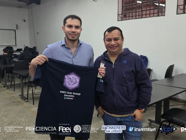 Two men pose in a classroom setting, with one holding up a black t-shirt featuring the AWS User Group Asunción logo. The image includes a banner of various organizational logos at the bottom.