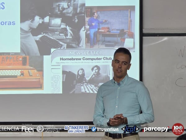 A man in a light blue shirt is giving a presentation in front of a projection screen displaying slides about 'AI Tinkerers' and the historical 'Homebrew Computer Club'.