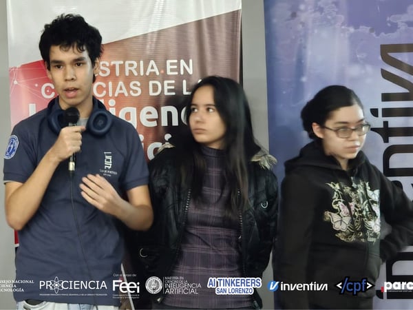 A group of three young people, including a young man speaking into a microphone, stand in front of event banners and a Paraguayan flag.