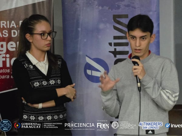 A man speaks into a microphone during a presentation while a woman stands beside him in front of event banners and logos.