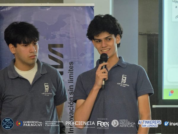 Two young men in matching blue polo shirts are presenting at an event, with one speaking into a microphone in front of a projection screen.