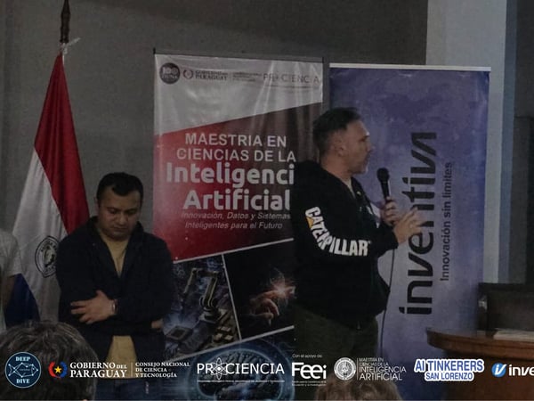 A man is giving a presentation in a room featuring promotional banners and a projection screen, while another man stands to the side.