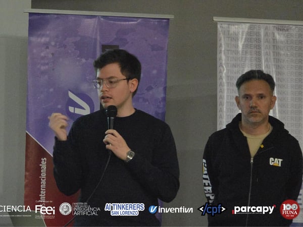 A man speaks into a microphone during a presentation, with another man standing beside him in front of event banners and a row of sponsor logos.