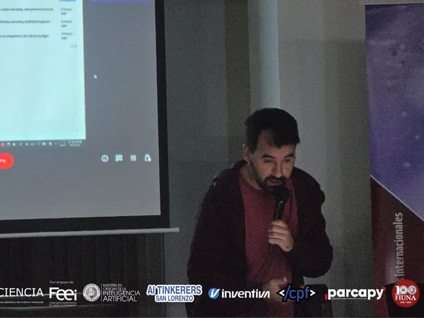 A man is speaking into a microphone while standing in front of a large projection screen displaying a data table during a presentation.