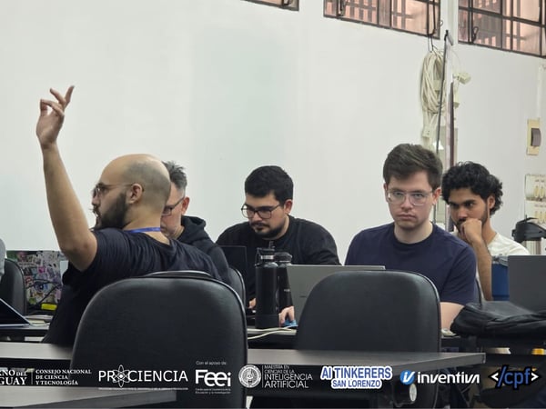 A group of men are seated at a table with laptops in what appears to be a workshop or meeting, with one man raising his hand to speak.