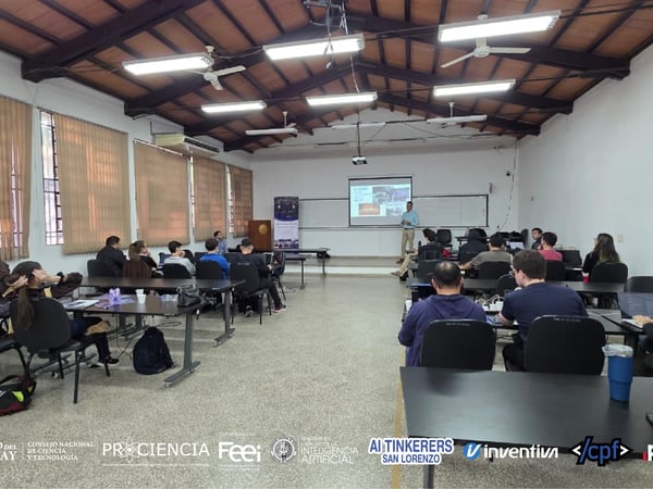 A group of people are attending a presentation in a classroom-style setting with a presenter standing at the front near a projector screen.